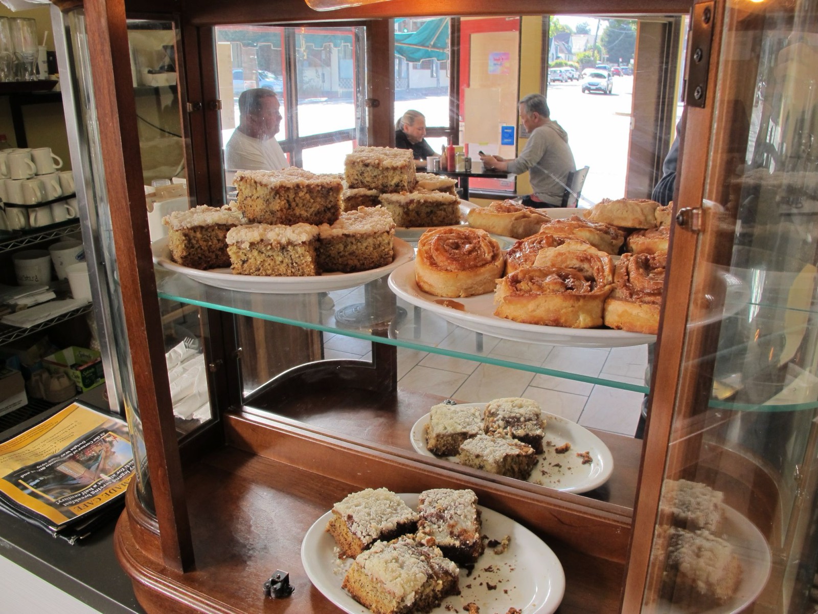 The Homemade Cafe pastry case filled with coffee cake and cinnamon rolls, with diners in the sunny dining room behind